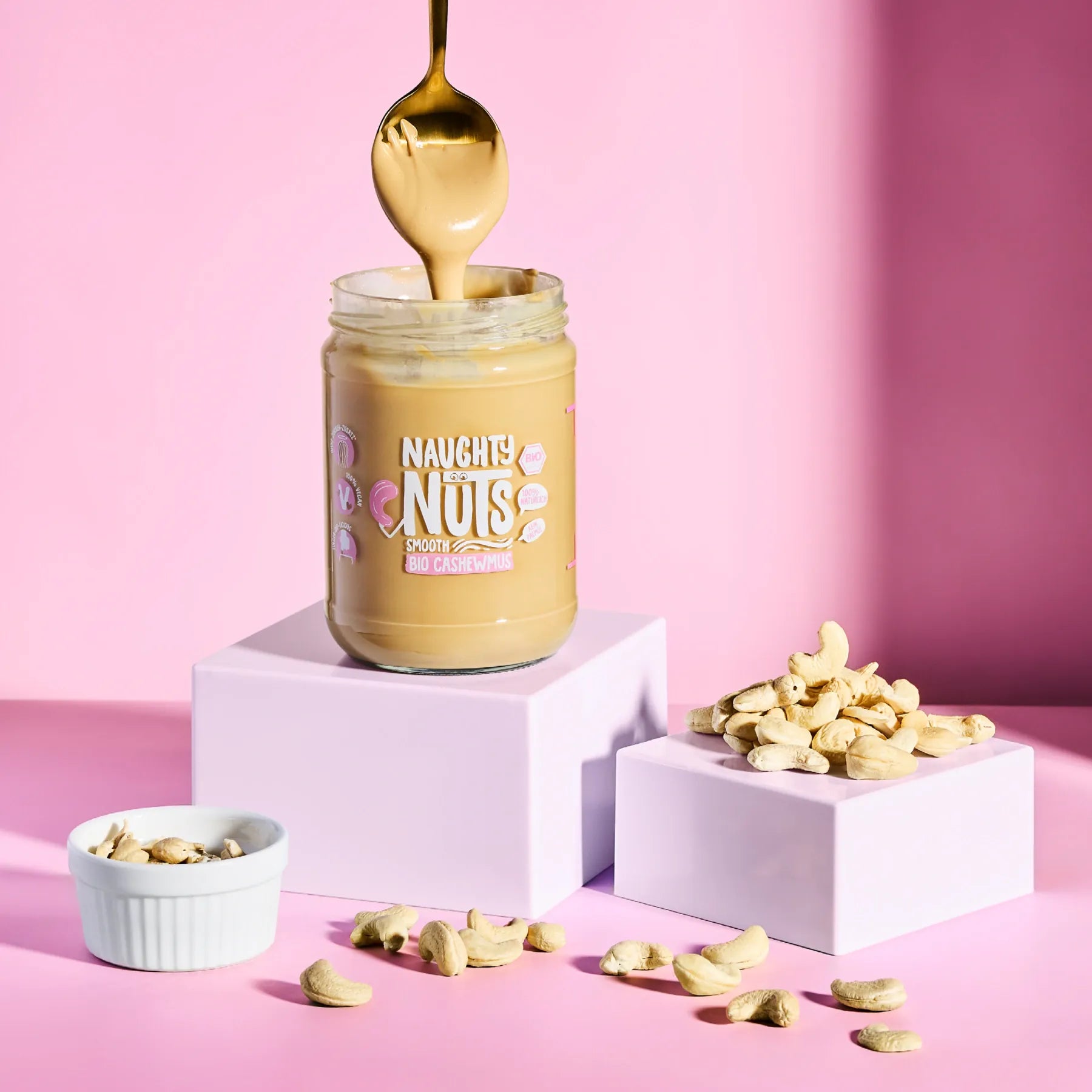 Bio Cashewmus Smooth im Glas mit Löffel und Cashewkernen auf rosa Hintergrund
