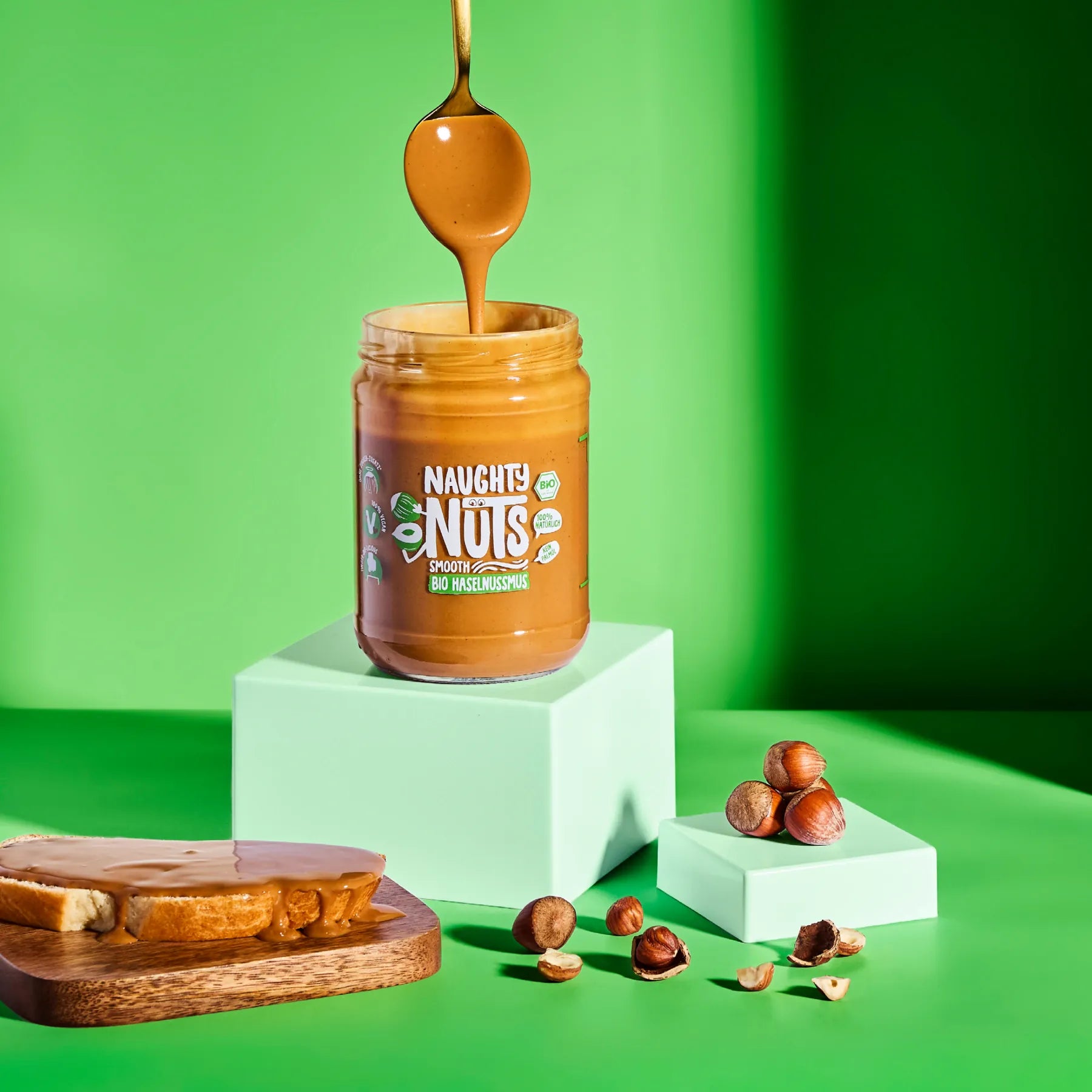 Bio Haselnussmus Smooth im Glas mit Löffel und bestrichener Brotscheibe auf grünem Hintergrund