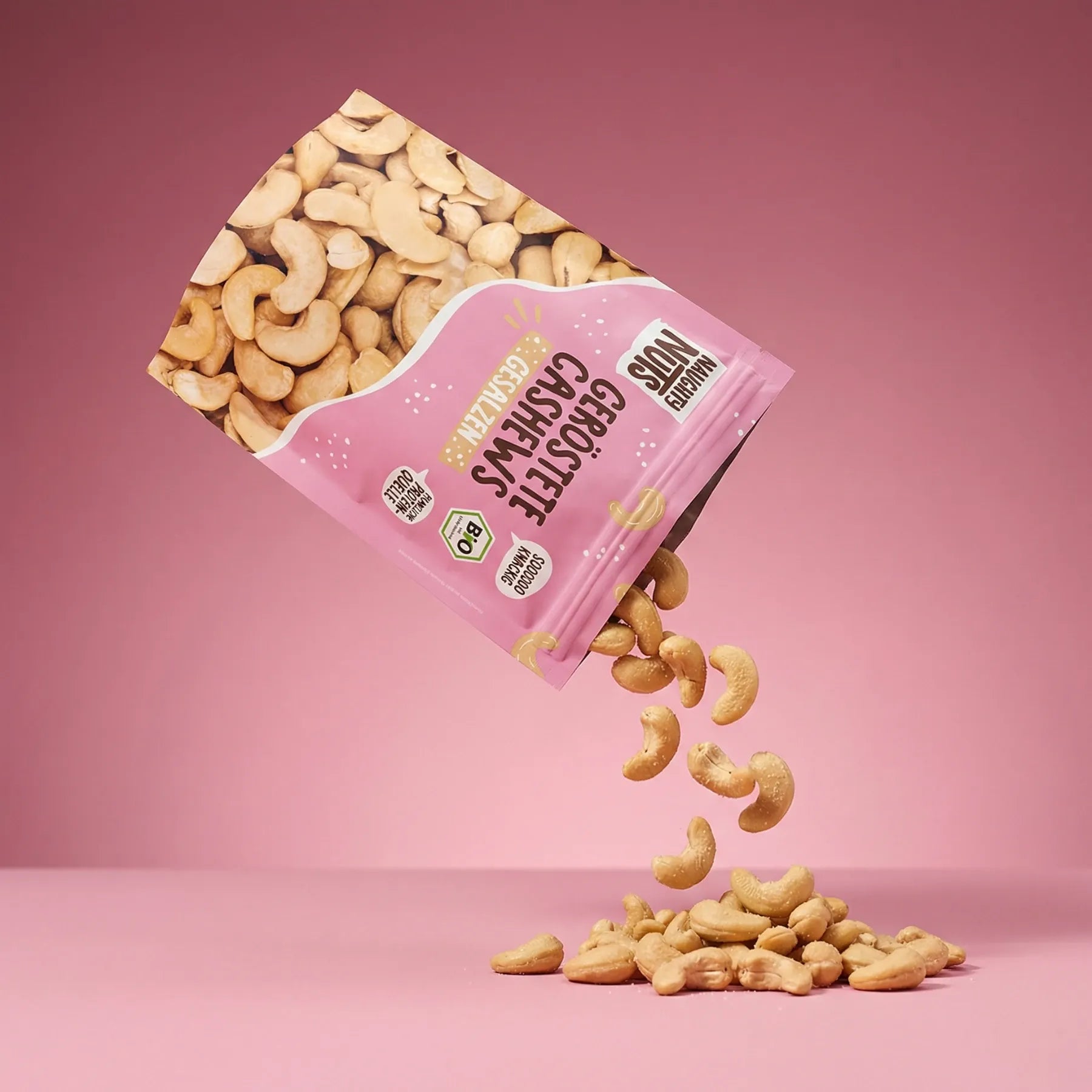 Geröstete Cashews gesalzen fallen aus einer rosa Verpackung auf einen rosa Hintergrund