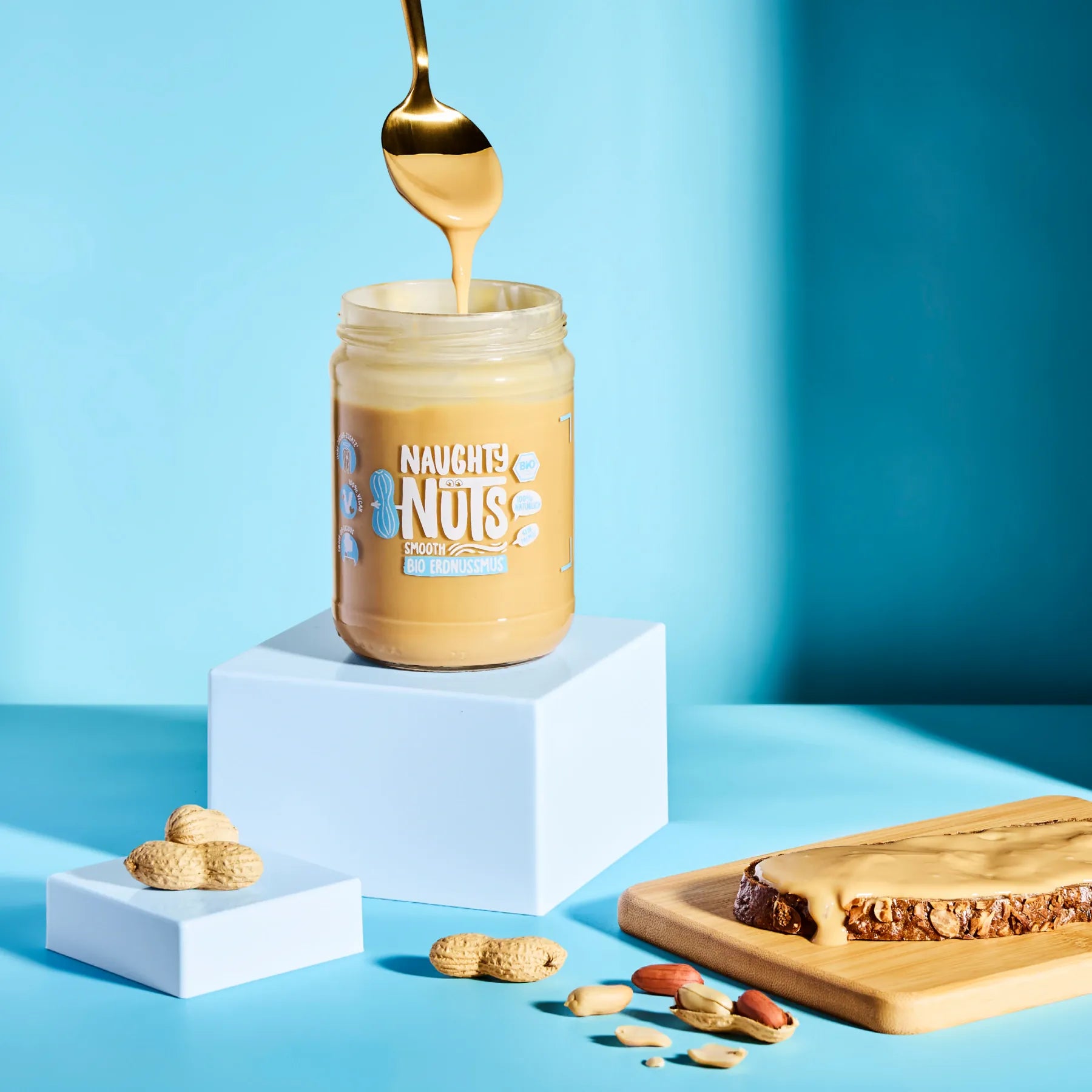 Bio Erdnussmus Smooth im Glas mit Löffel auf blauem Hintergrund und einer Scheibe Brot