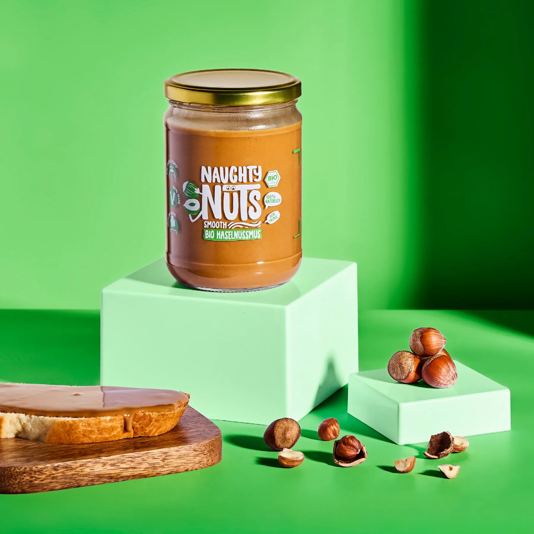 Bio Haselnussmus Smooth im Glas auf einem Podest mit Haselnüssen und einer Scheibe Brot