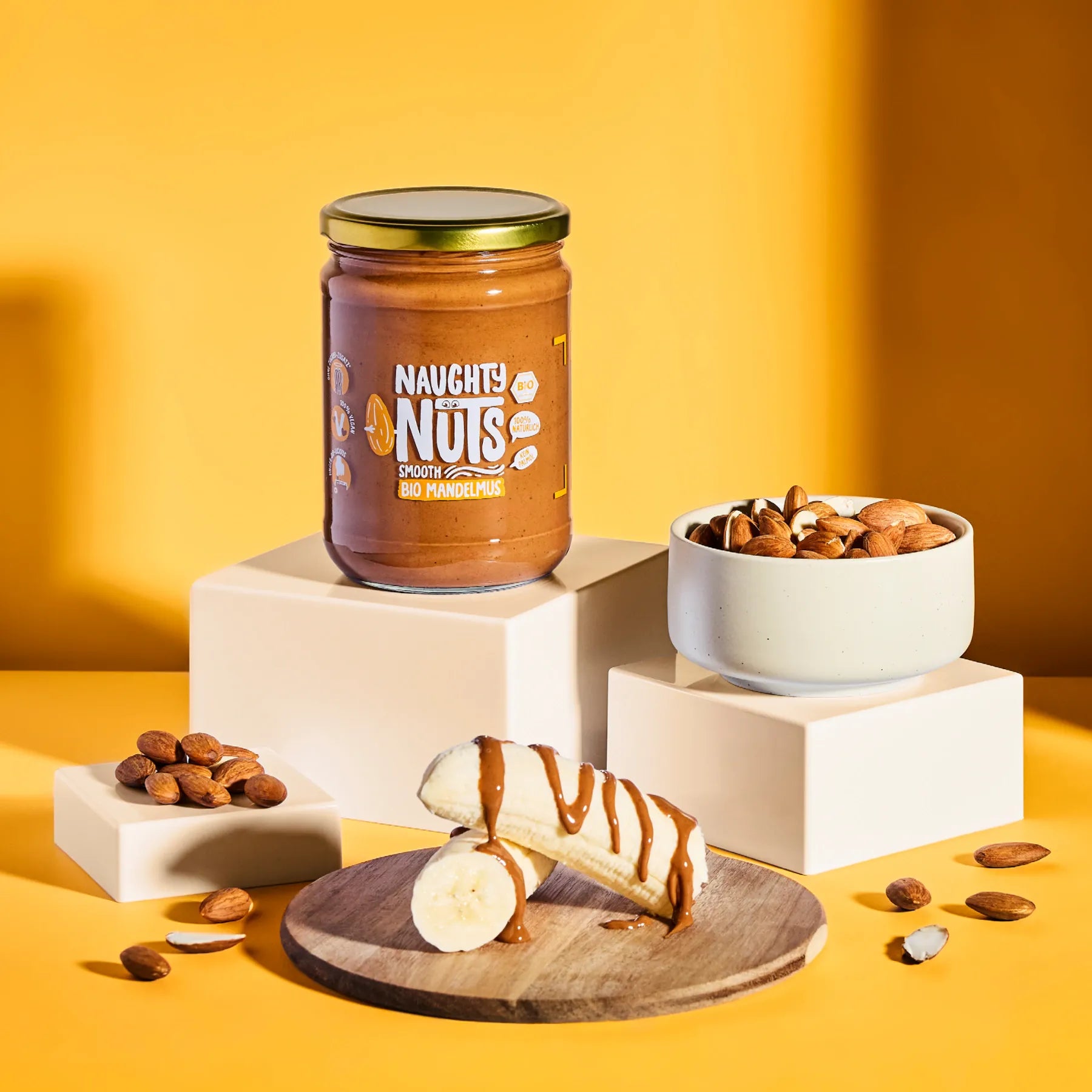 Bio Mandelmus Smooth im Glas neben einer Schale mit Mandeln und Bananenstücken mit Mandelmus