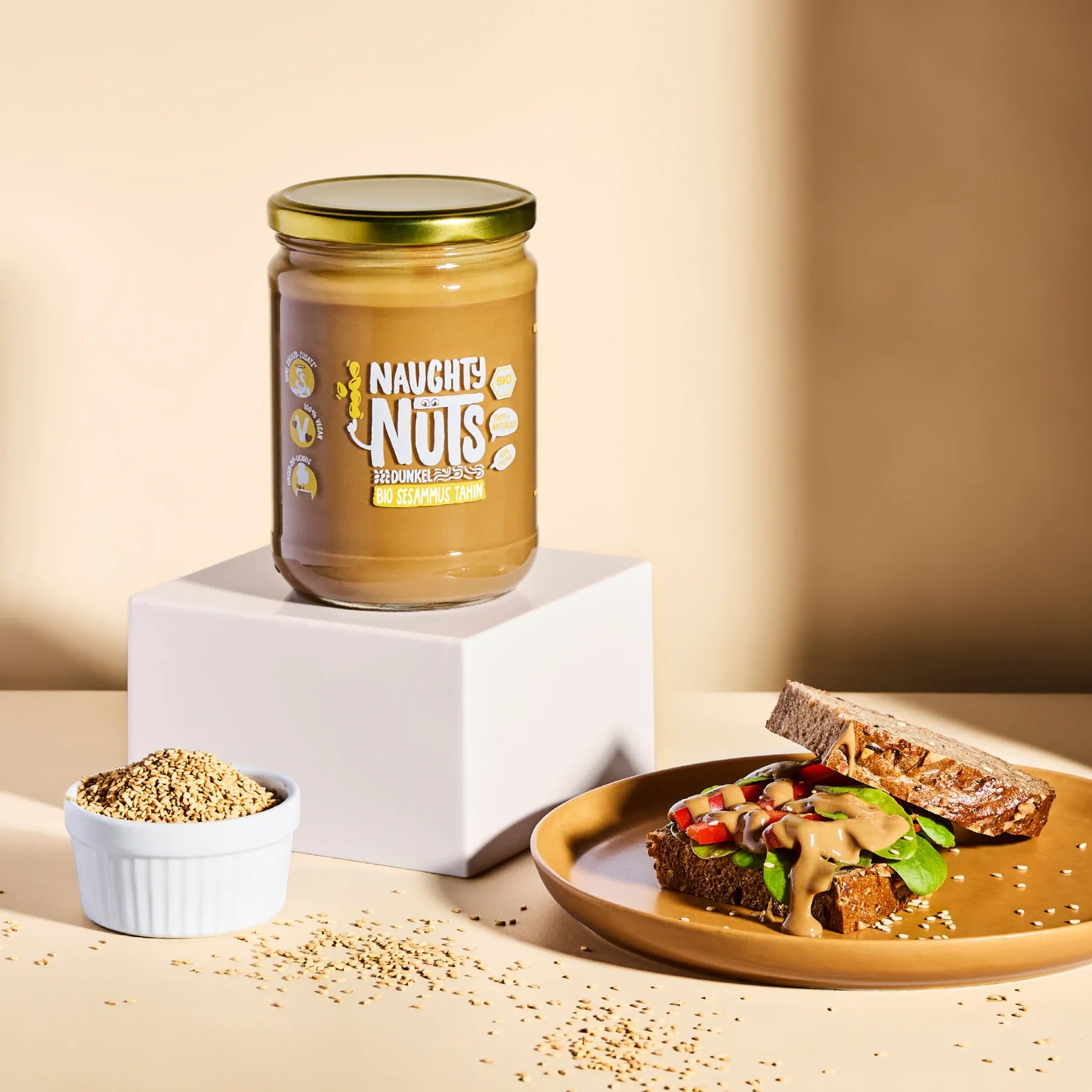 Bio Sesammus Tahin dunkel im Glas auf einem Podest neben einer Scheibe Brot mit Tahin und einer Schale Sesam