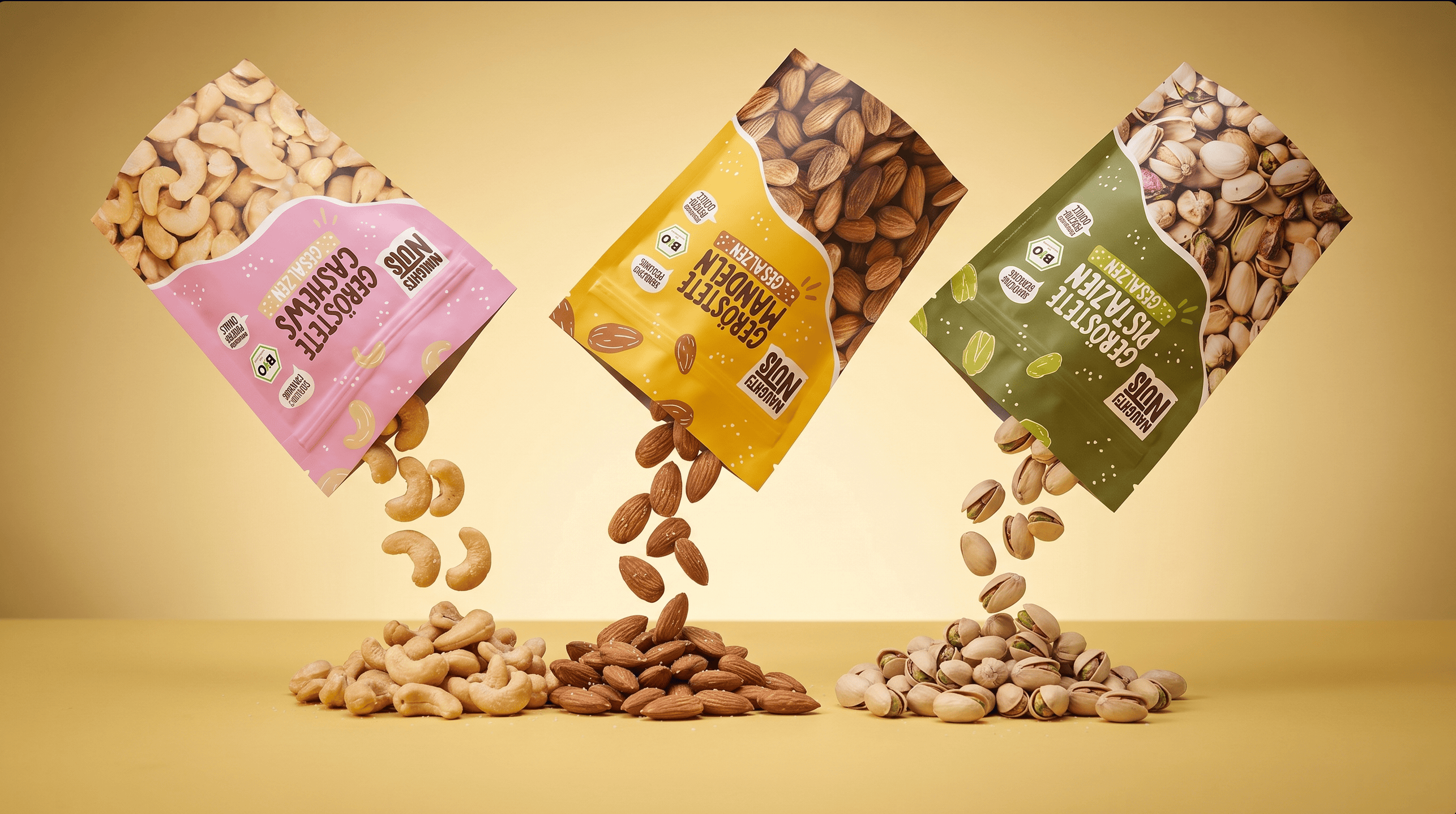 Drei Packungen Naughty Nuts: geröstete Cashews, Mandeln und Pistazien, die auf einen gelben Tisch fallen